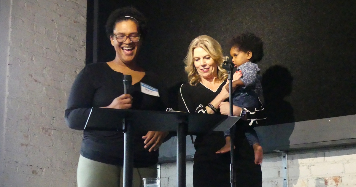 Celebrating Brittni's Family Home: Brittni speaks into a microphone while WLNS 6 anchor Sheri Jones holds Brittni's youngest child.