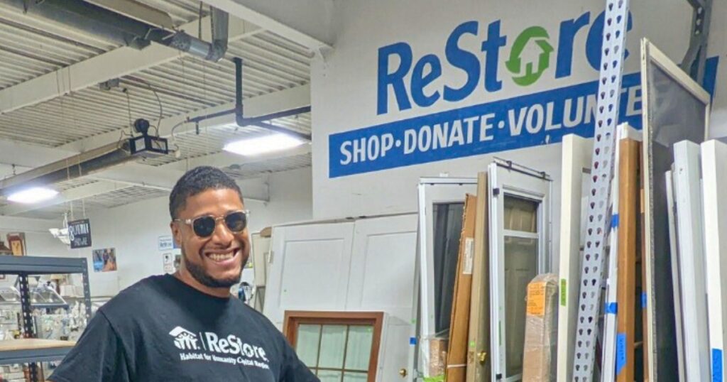 Josh, ReStore employee, smiles for a photo in the front of the ReStore logo for Secondhand September.